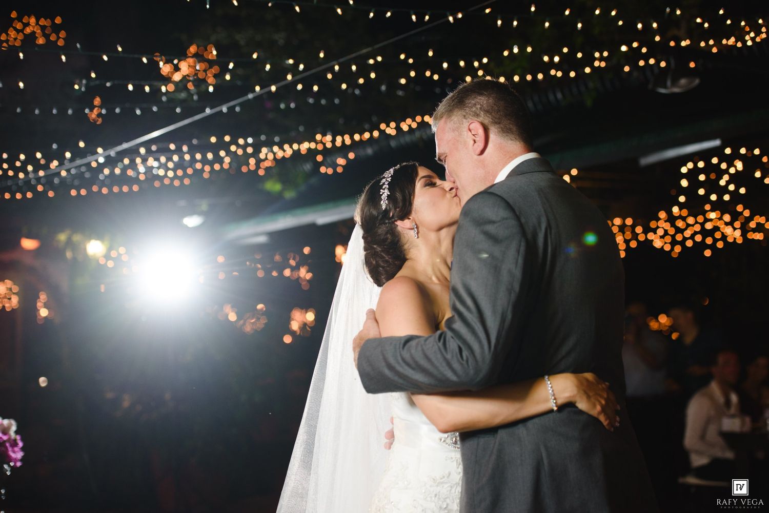 Newlyweds share romantic kiss on dance floor surrounded by glowing string lights at wedding reception.