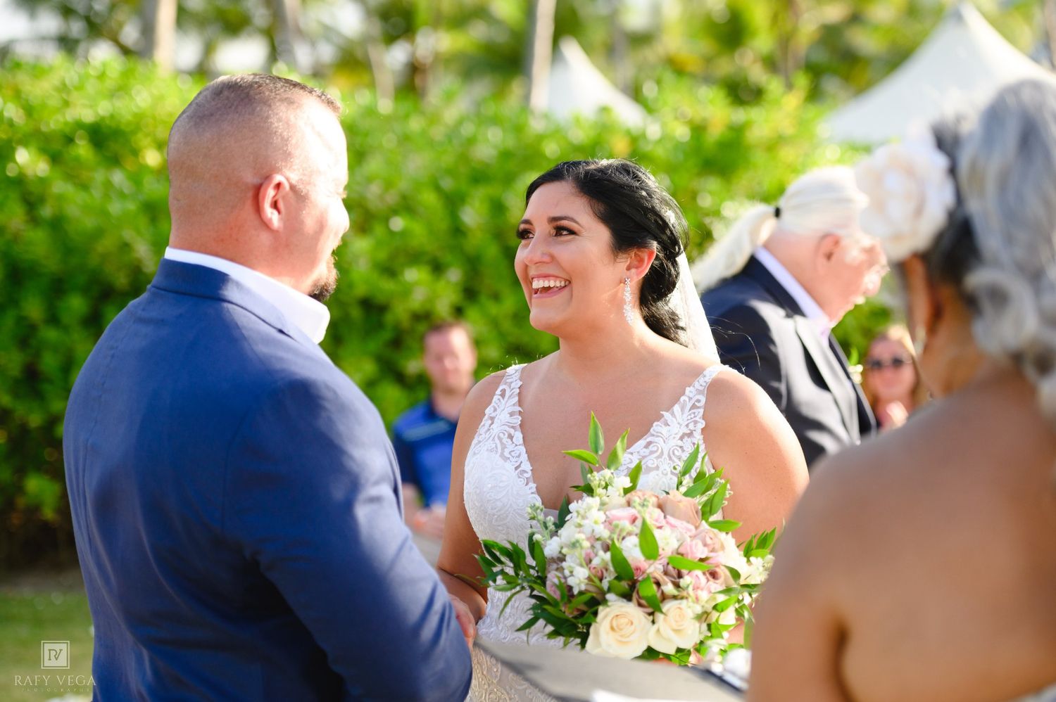 Bride smiling joyfully at groom during outdoor beach wedding ceremony, holding white and pink floral bouquet.
