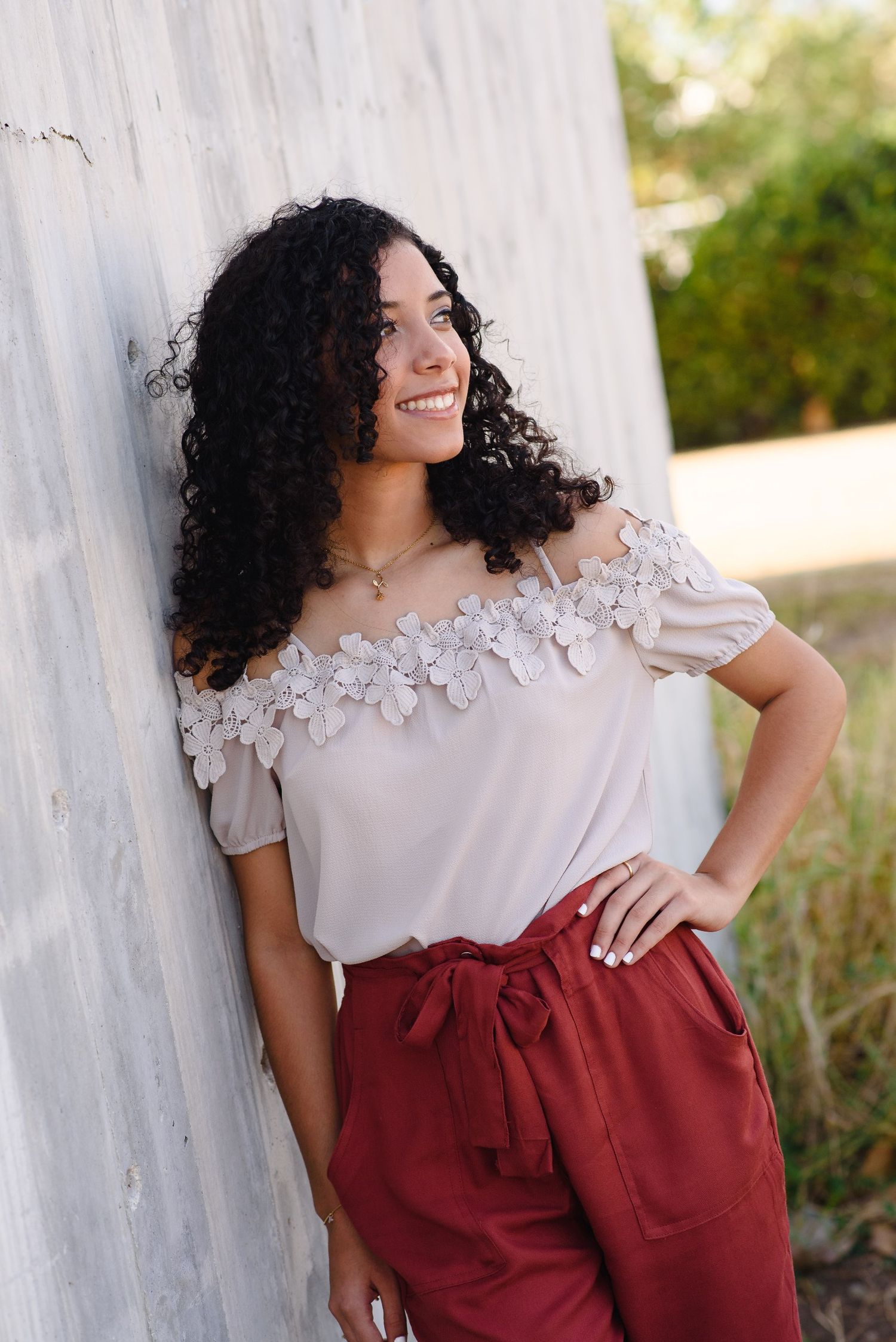 Young woman with curly hair smiling, wearing off-shoulder floral top and red pants, leaning against a wall.