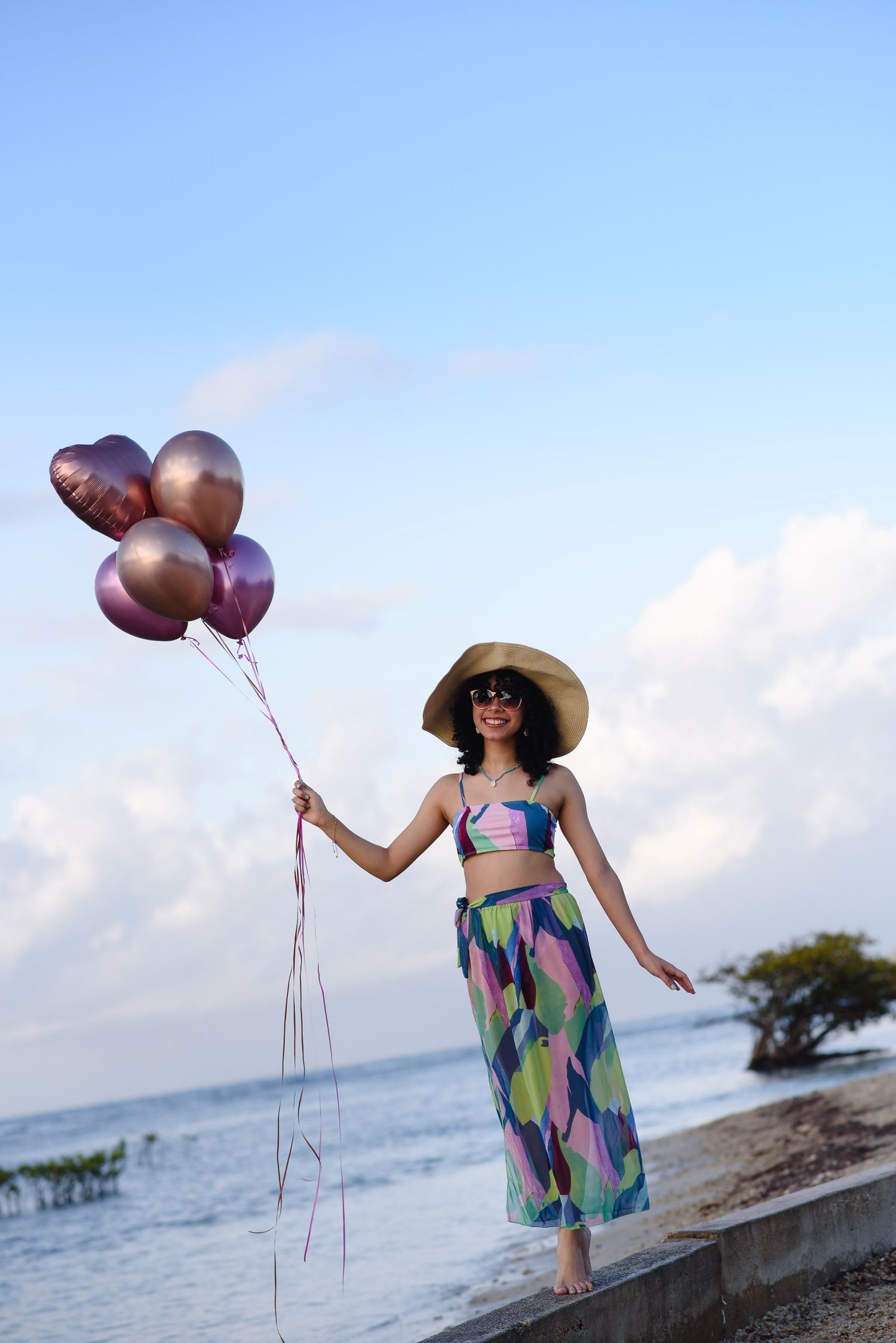 Woman in colorful outfit and sun hat holds pink heart balloons by the ocean on a sunny day.