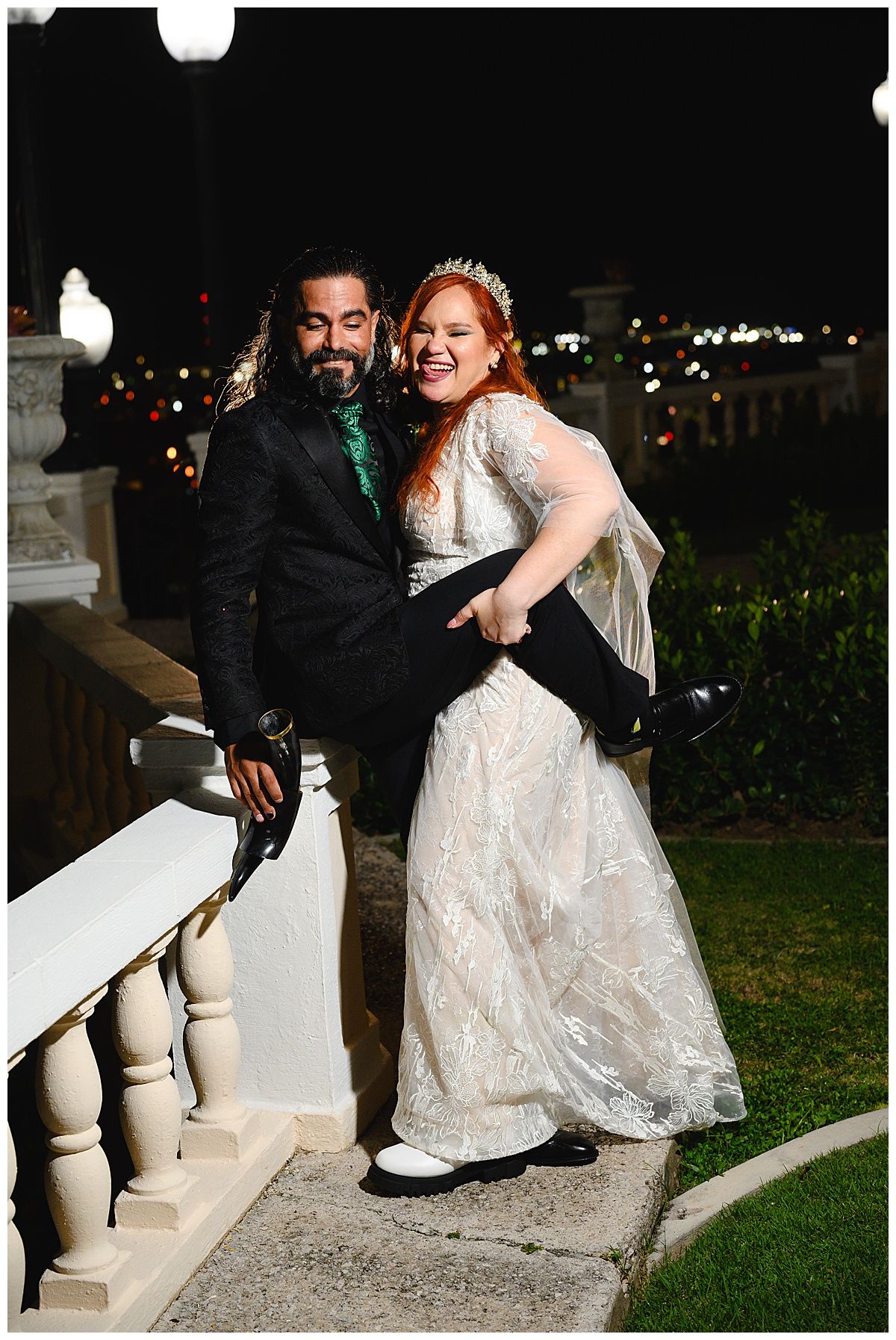 Groom lifts bride in white lace gown at night wedding, posing by white balustrade with city lights behind.