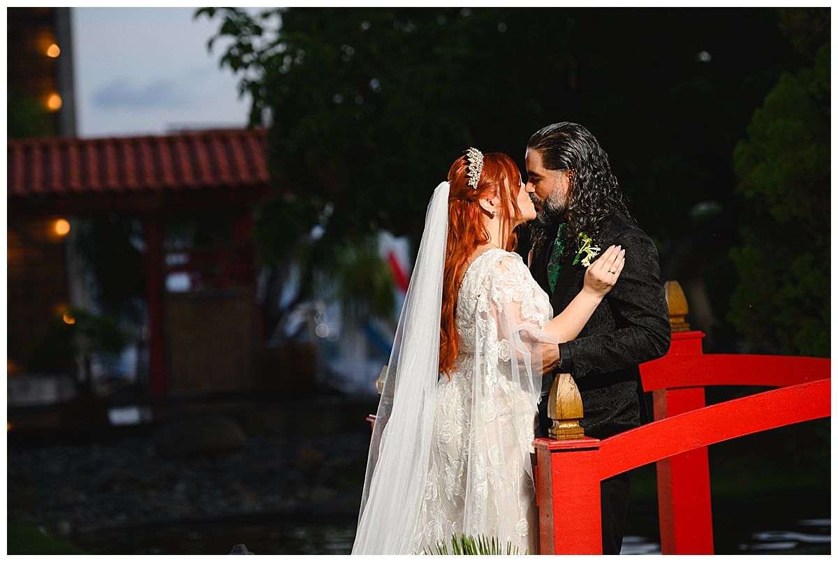 Bride and groom share a romantic kiss on a red bridge, bride wearing lace gown and veil.