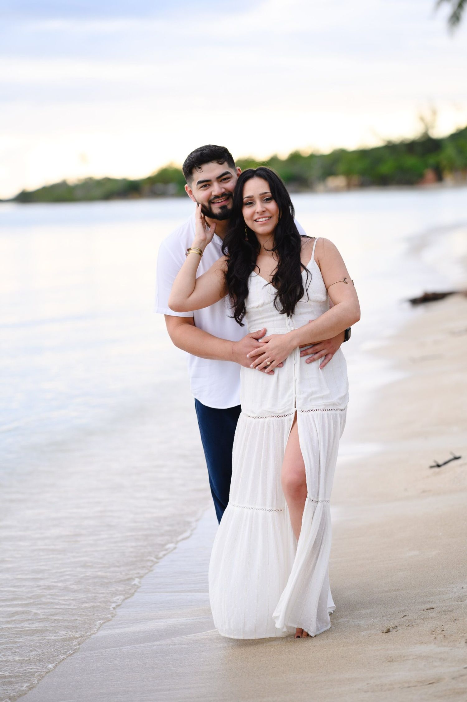 Couple embracing on a sandy beach at sunset, woman in white maxi dress, man in white shirt and navy pants.
