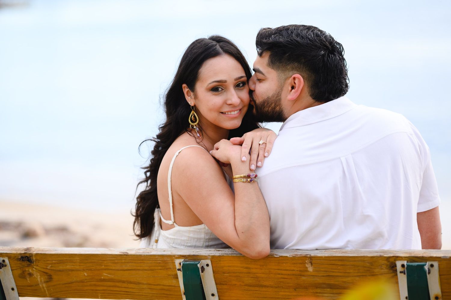 Romantic couple on a beach bench, man kissing woman's cheek as she smiles, ocean visible in the background.