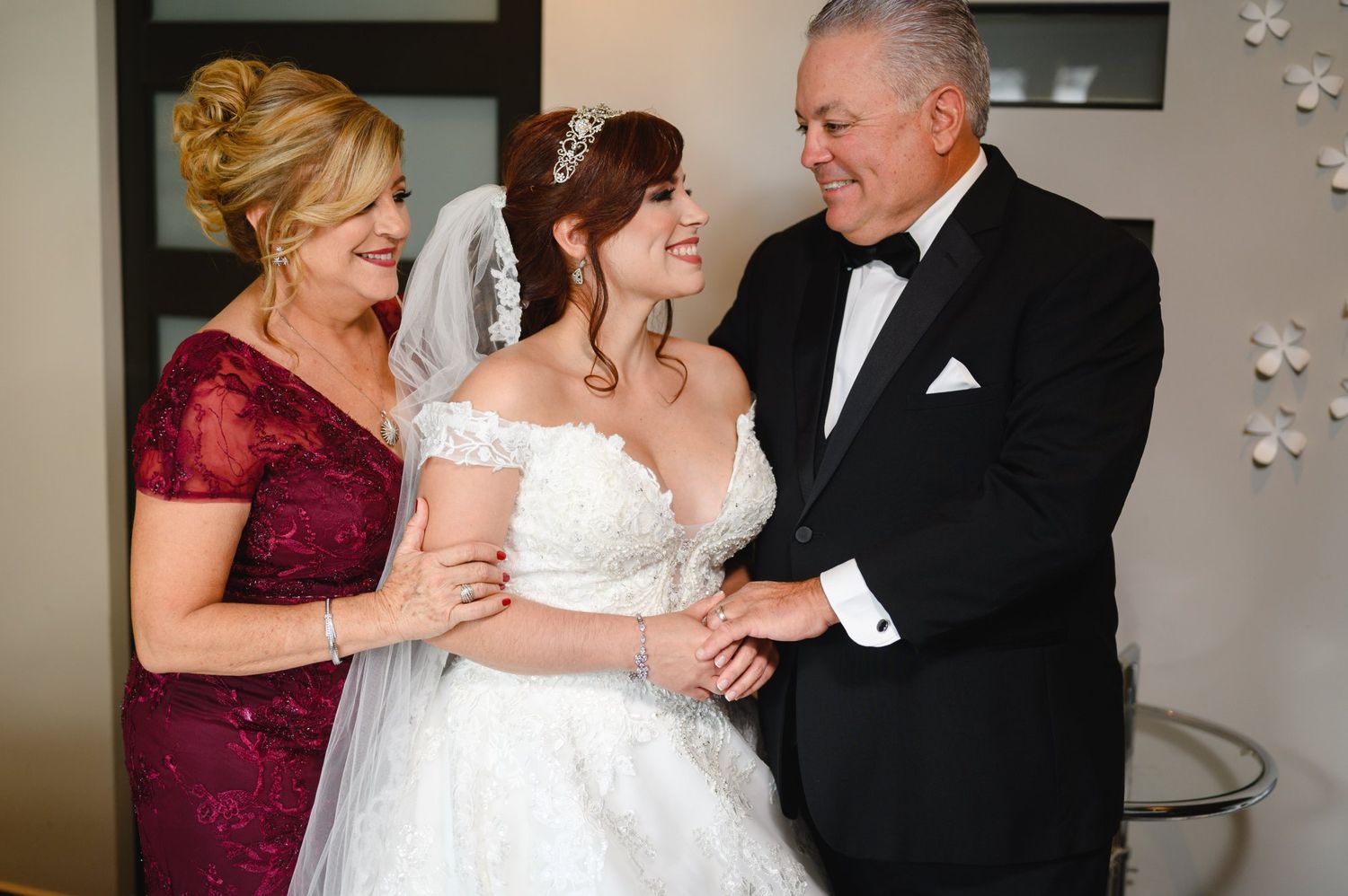Bride in white lace gown smiles with parents in formal attire at wedding celebration.