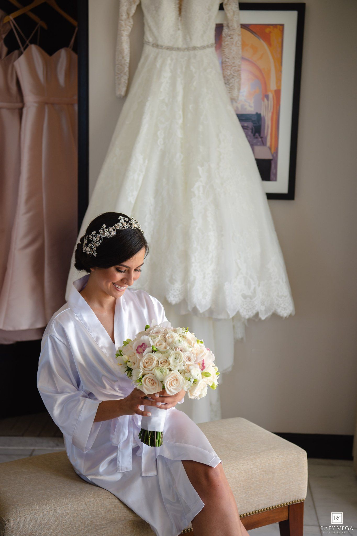 Bride in white robe holding floral bouquet with wedding gown hanging in background.