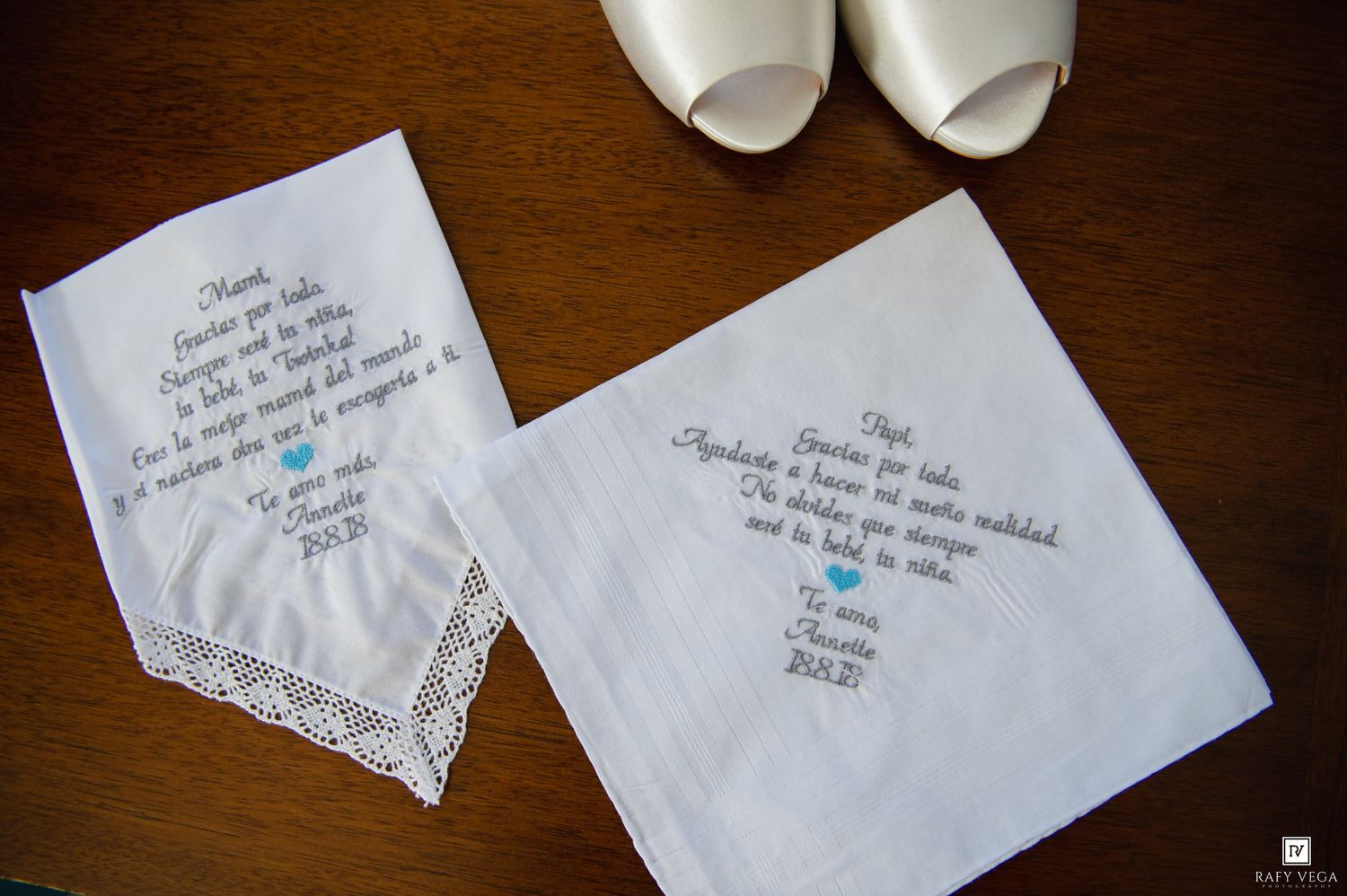 Two embroidered wedding handkerchiefs with heartfelt messages displayed on a wooden surface near white bridal shoes.