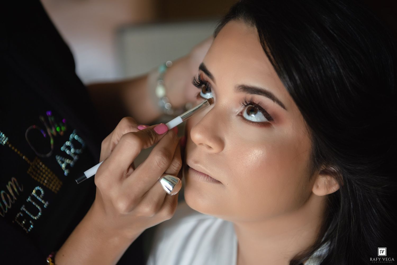 Professional makeup artist applying eye makeup on a young woman with dark hair and glam beauty look.