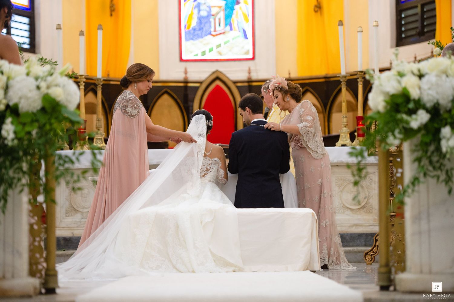 Bride and groom exchanging vows at a church altar with bridesmaids in pink gowns and white floral arrangements.