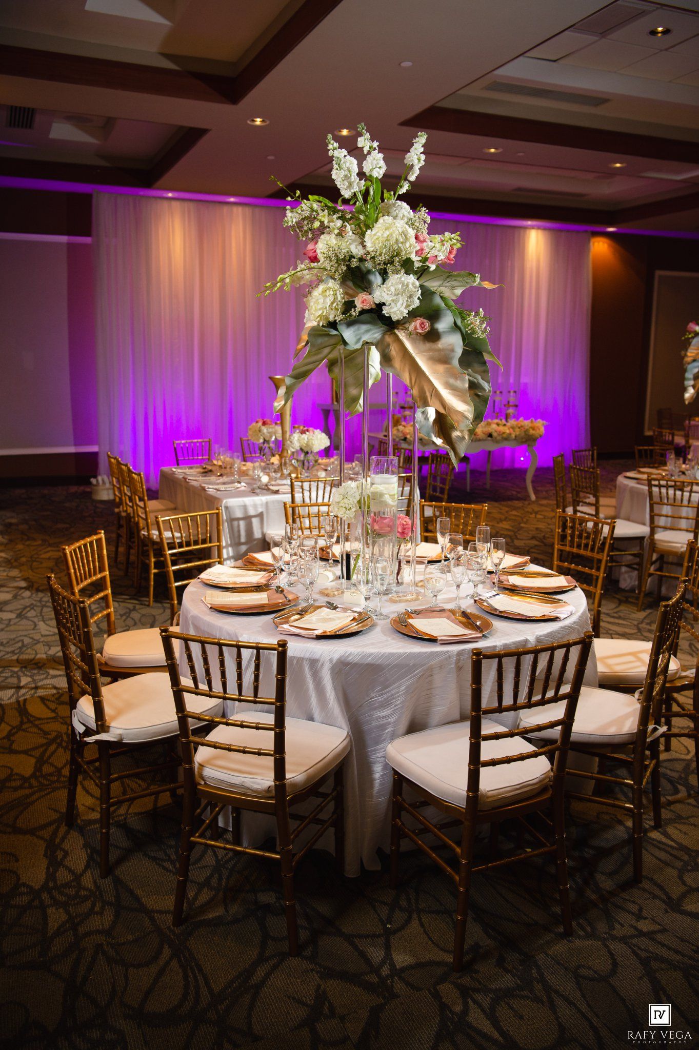 Elegant wedding reception table with tall floral centerpiece, gold chiavari chairs, and purple ambient lighting in ballroom.