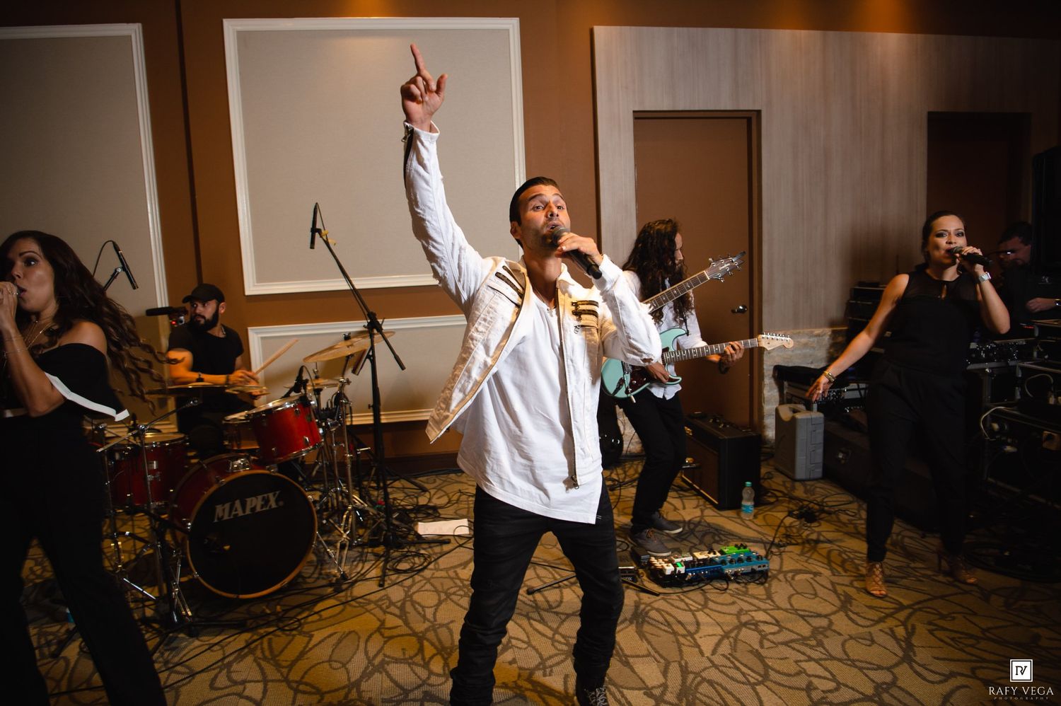 Live band performance with energetic male singer pointing up, guitarist and drummer on stage in elegant venue.