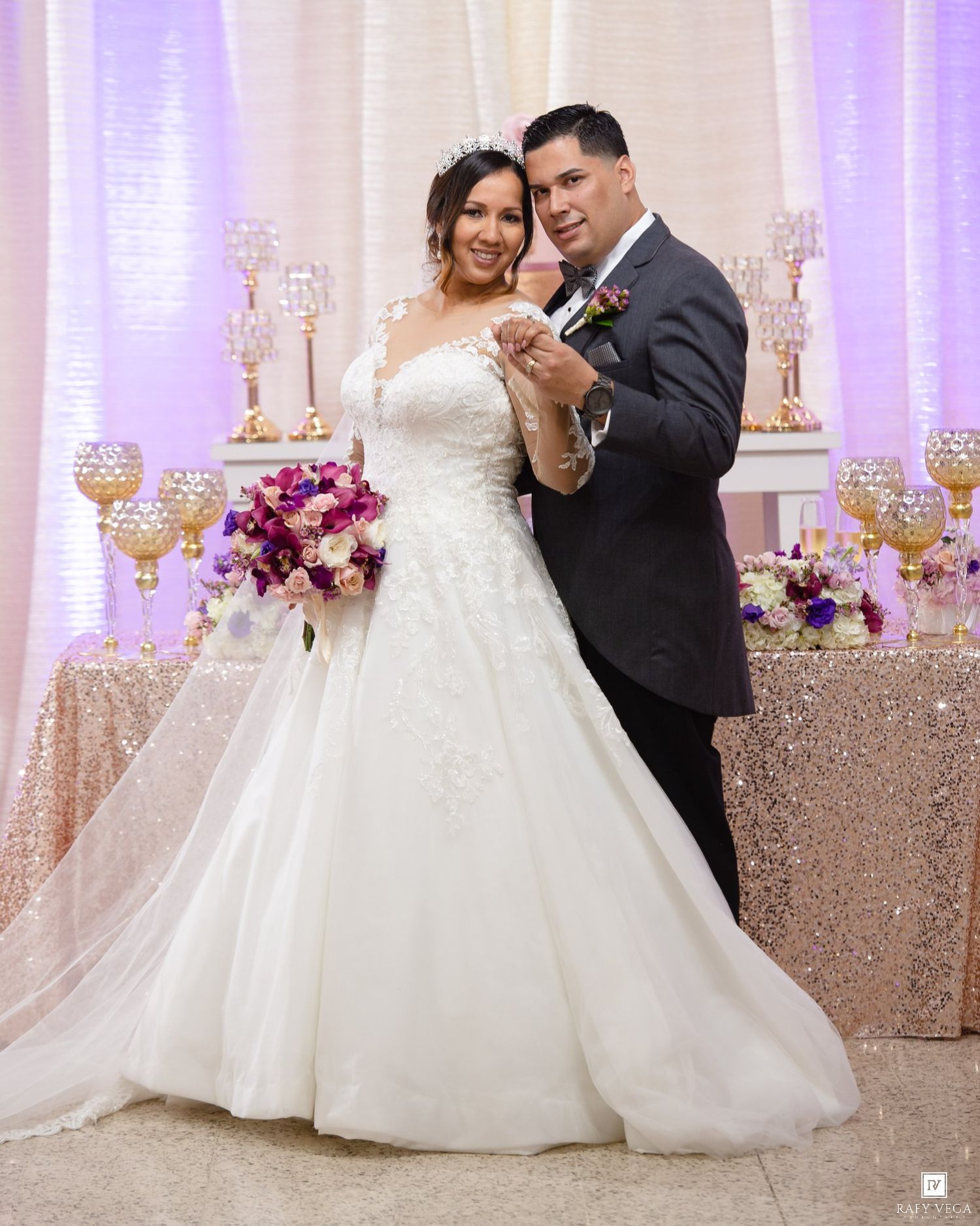 Bride in white ball gown and groom in gray tuxedo pose together at elegant wedding reception with purple backdrop.