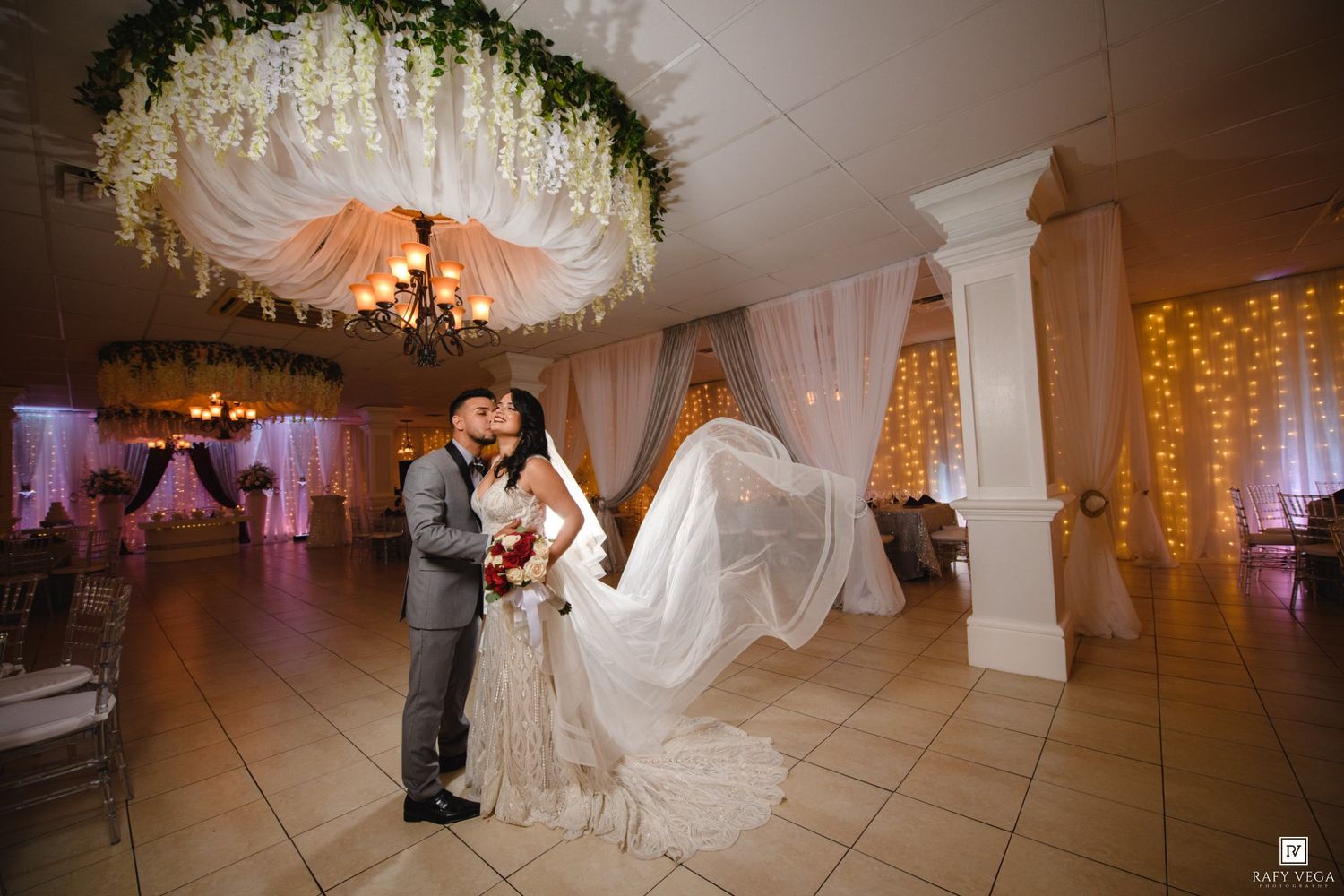 Bride and groom share a romantic kiss in an elegantly decorated wedding reception hall with floral ceiling decor.