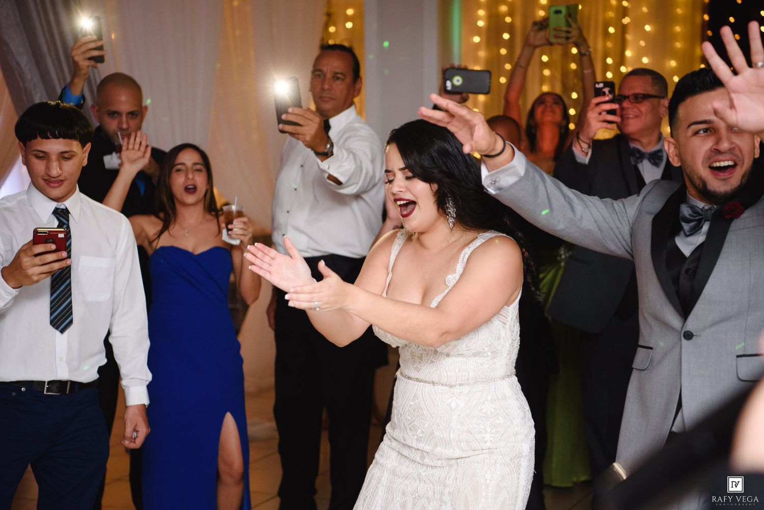 Bride and guests celebrate joyfully on wedding reception dance floor with phones raised.