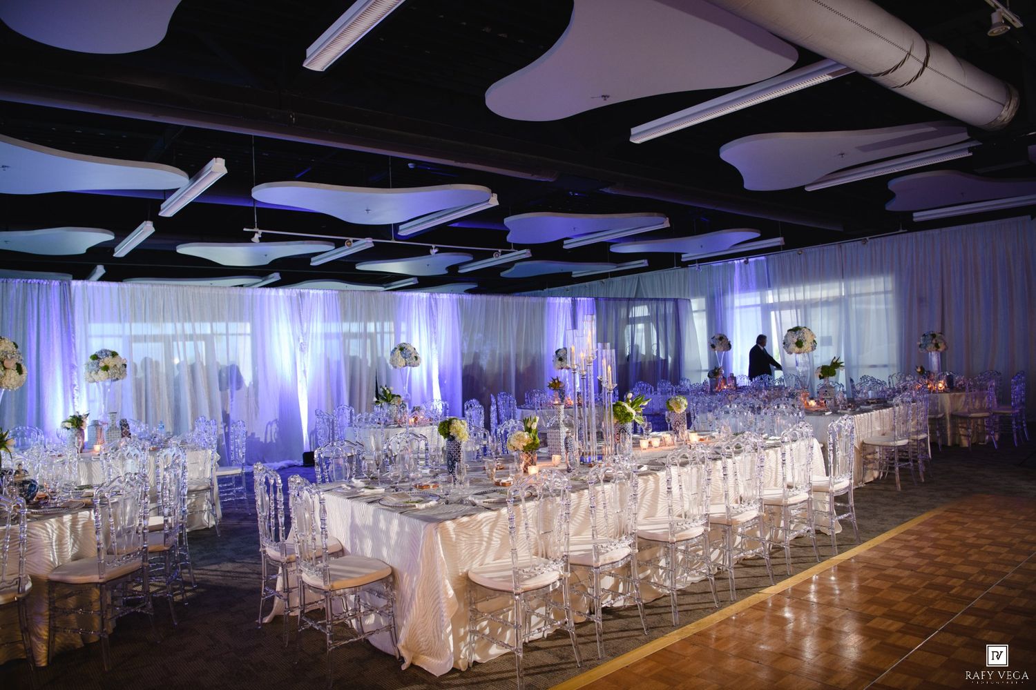 Elegant wedding reception hall with white draping, crystal chairs, long banquet tables, and purple uplighting.
