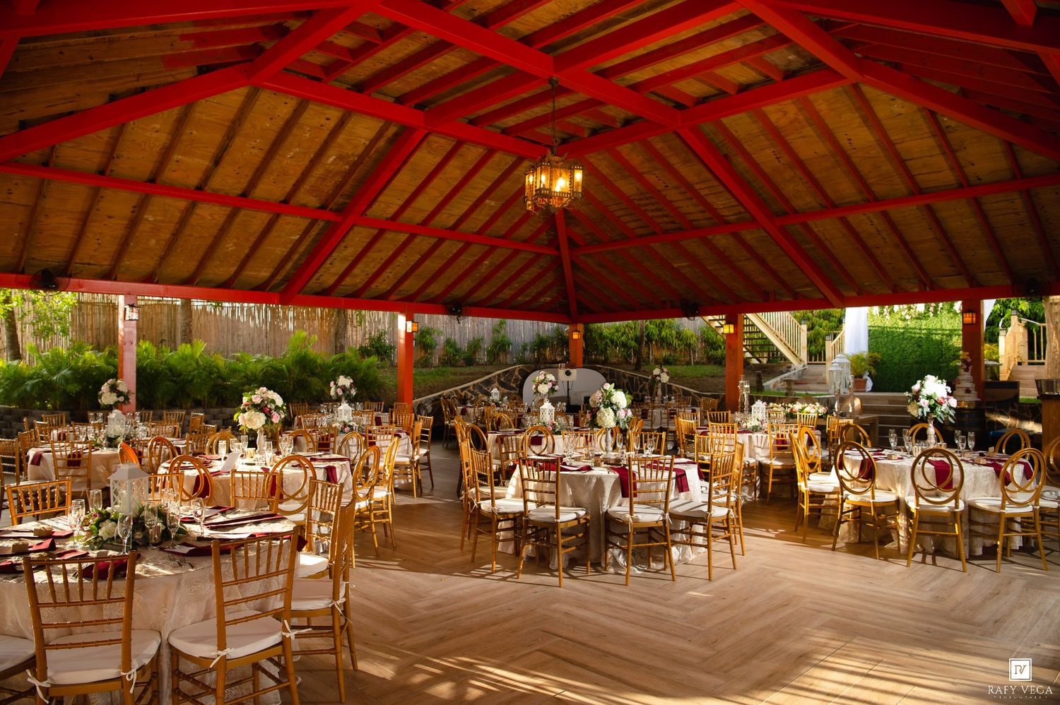 Elegant outdoor wedding reception under a red-roofed pavilion with gold chiavari chairs and floral centerpieces.