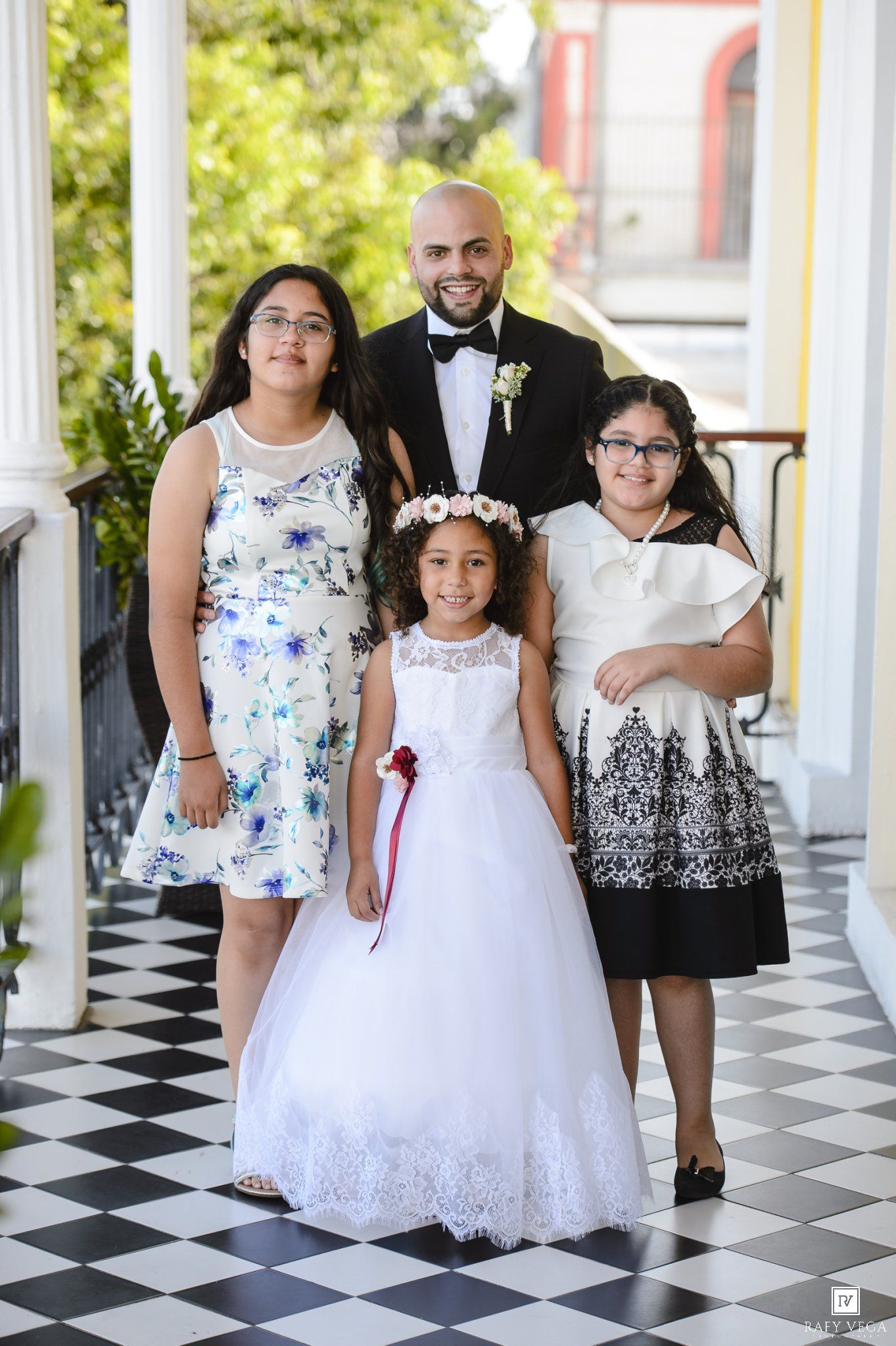 Man in tuxedo poses with three girls in formal dresses on a checkered floor at a wedding celebration.