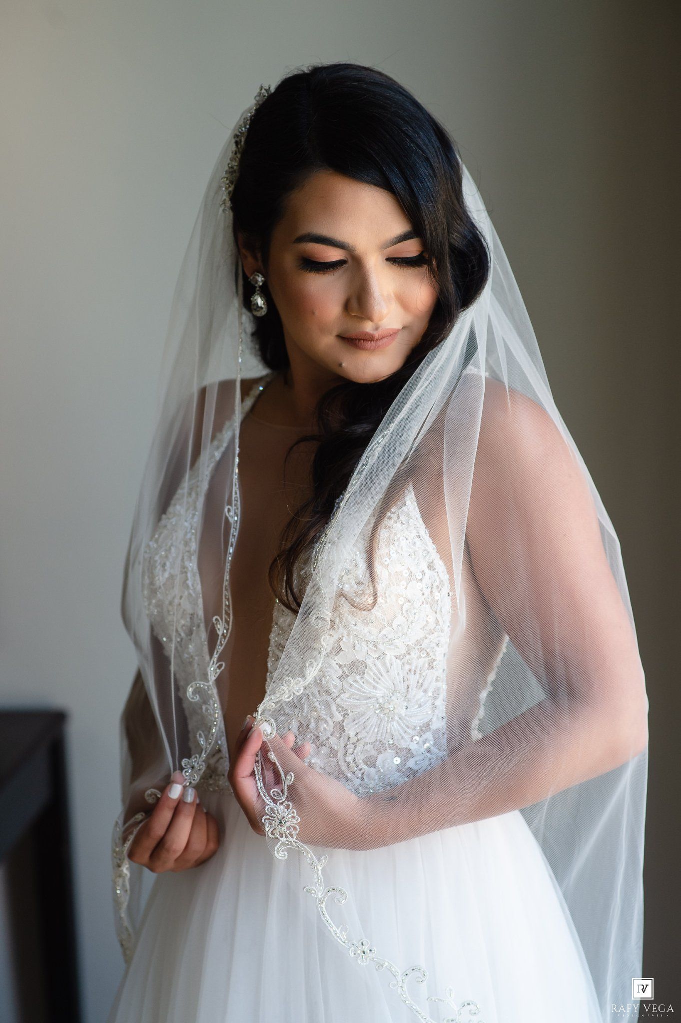 Elegant bride in lace wedding dress and long veil looks down softly while holding her gown indoors.