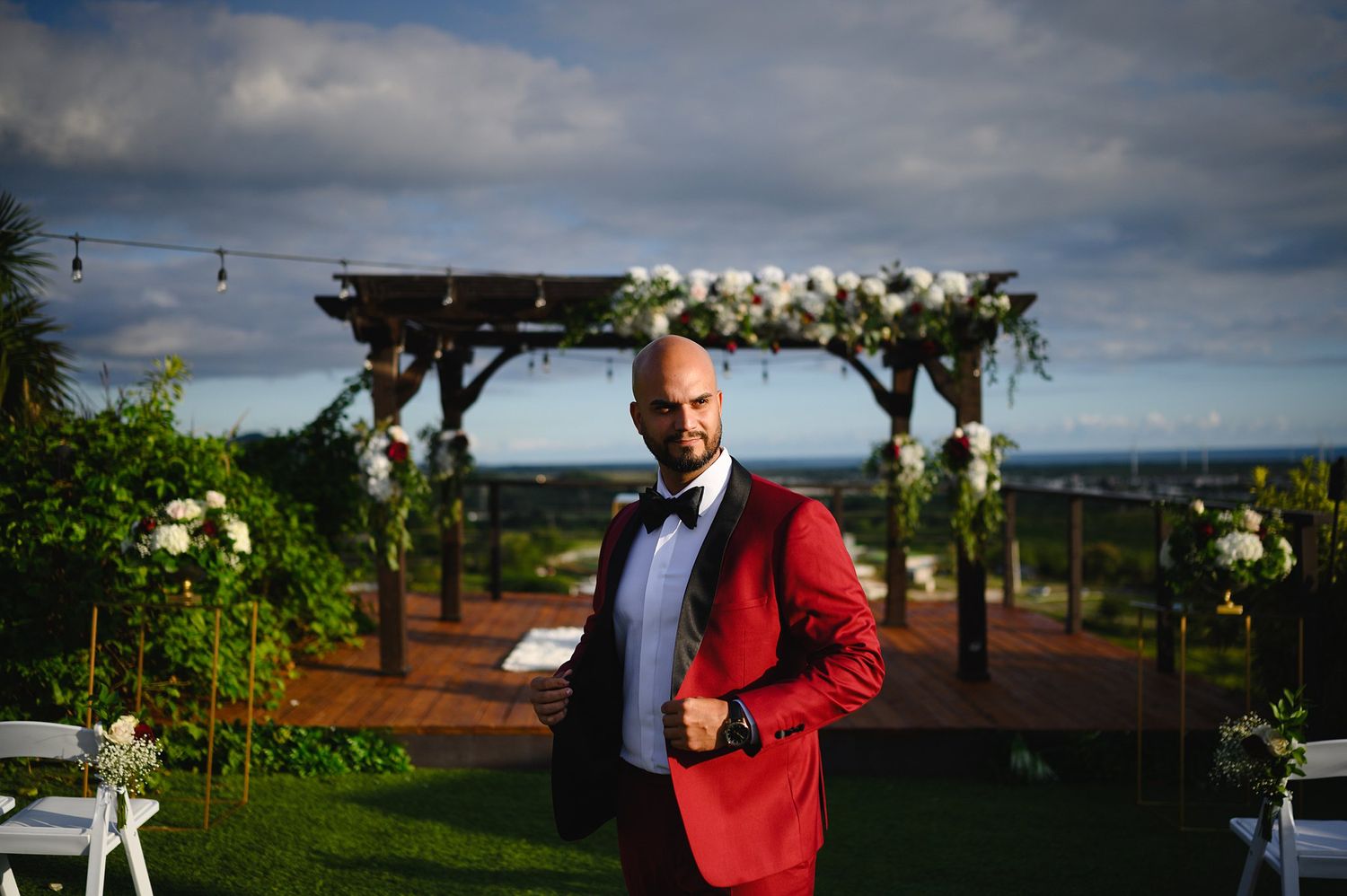 Groom in red tuxedo jacket stands before floral arch at outdoor wedding ceremony on lush green lawn.