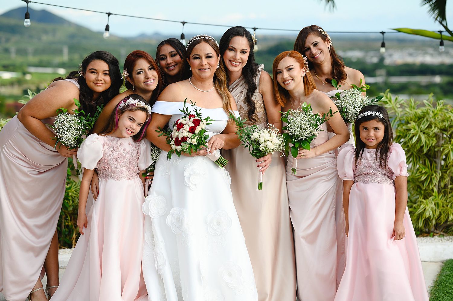 Bride in white gown poses with bridesmaids in blush pink dresses and flower girls holding bouquets outdoors.