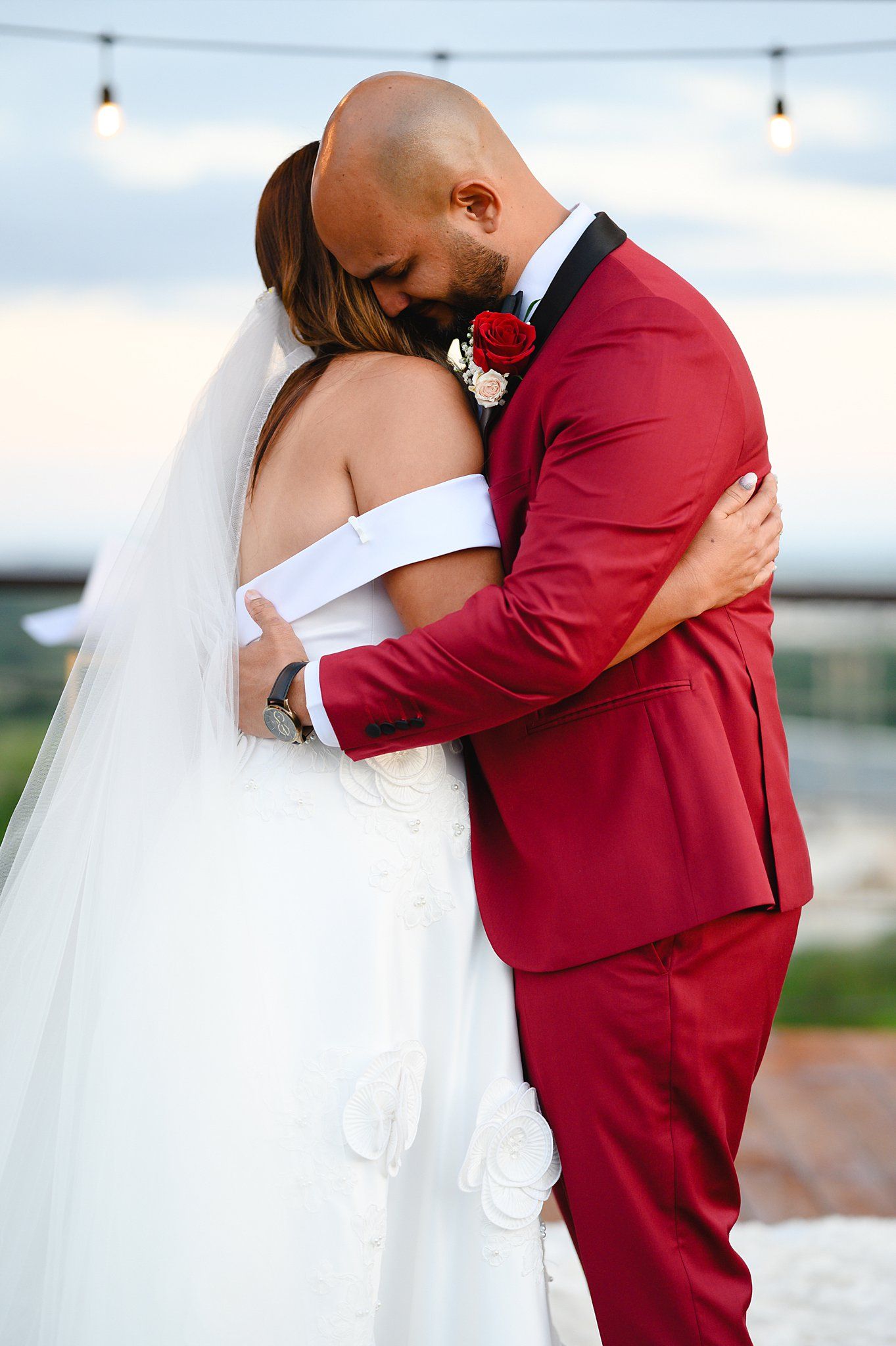 Bride and groom share an intimate embrace, she in a white gown with veil, he in a bold red suit with boutonniere.