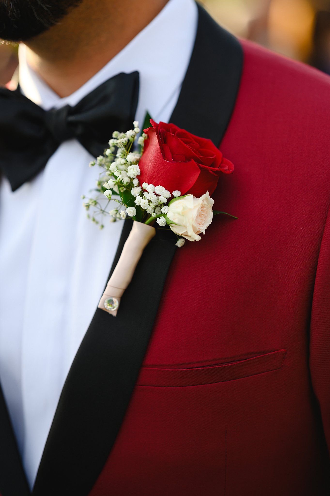 Man in red tuxedo with black lapels and bow tie wearing a boutonniere of red and white roses with baby's breath.