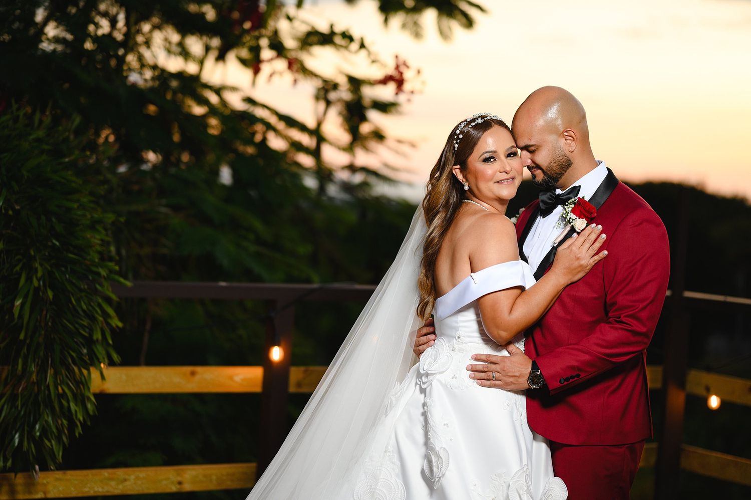 Bride in white gown and groom in red suit embrace at sunset during romantic outdoor wedding ceremony.