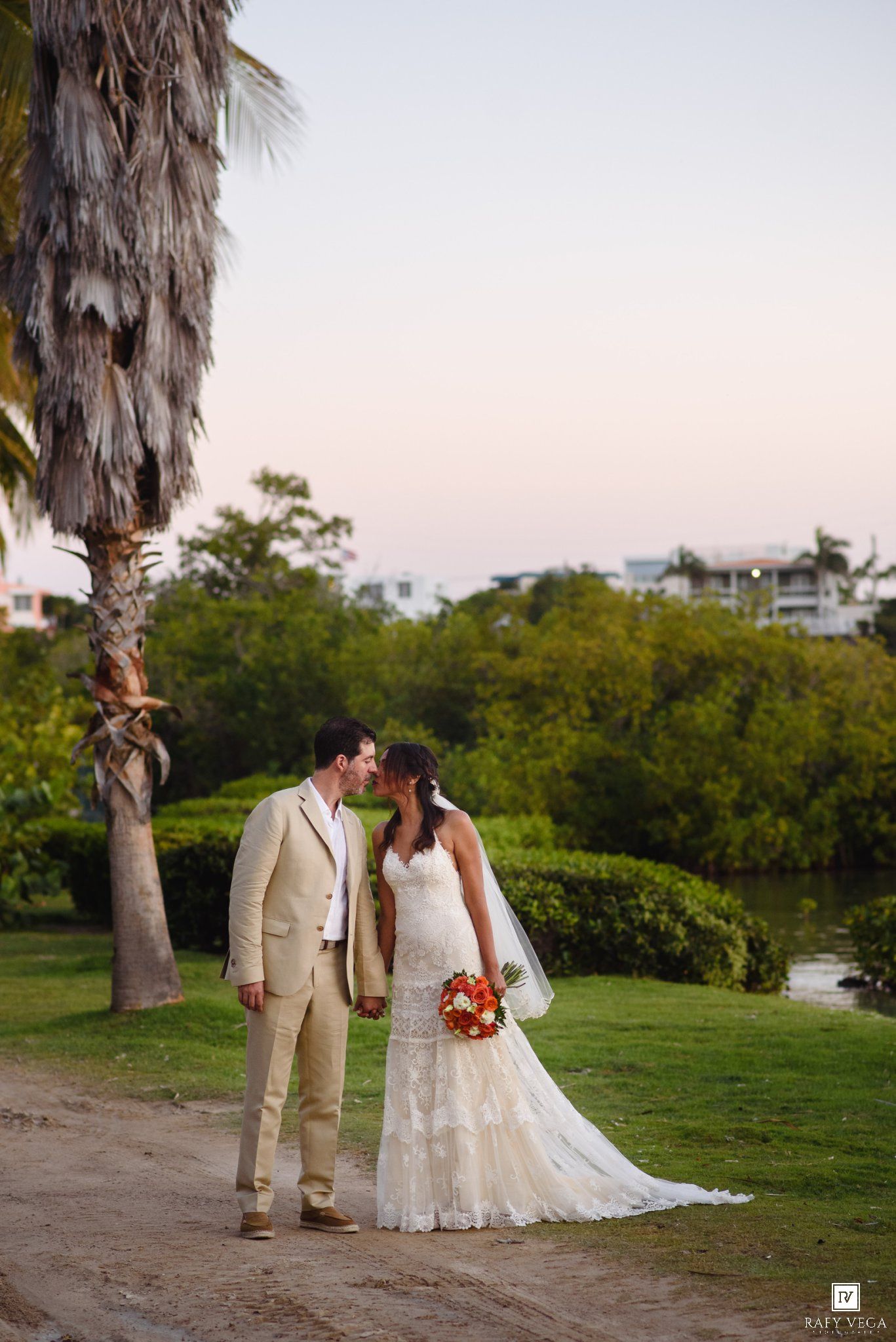Bride and groom share a kiss on sandy path surrounded by lush greenery at sunset.