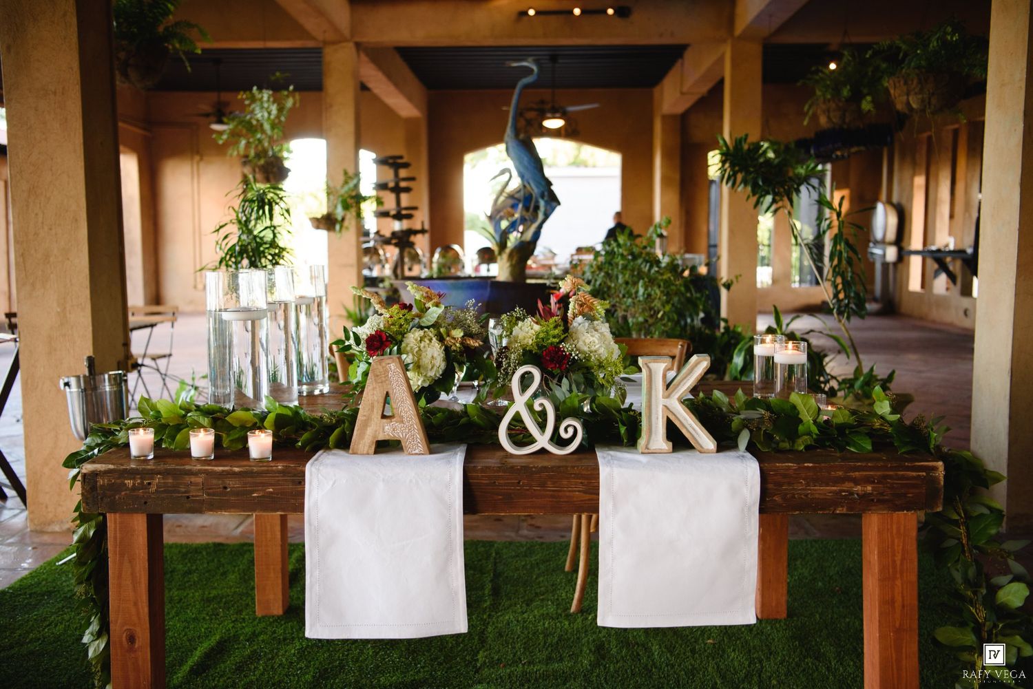 Rustic wooden sweetheart table decorated with greenery, candles, and monogram letters A&K for an elegant wedding reception.