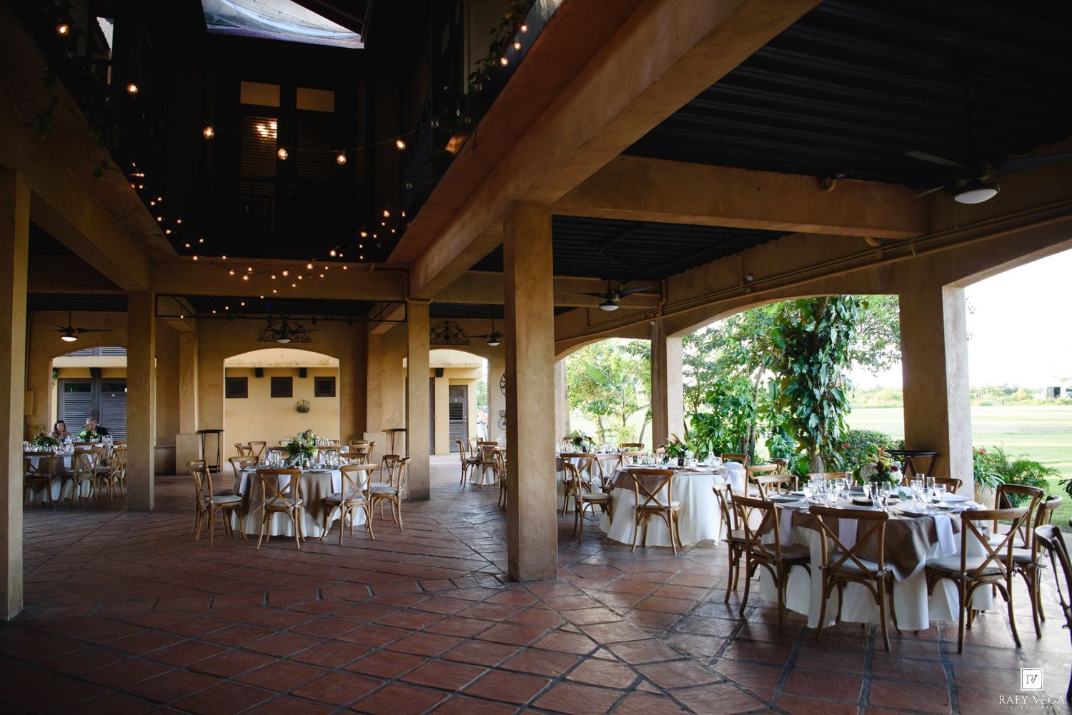 Spacious outdoor wedding venue with string lights, terracotta floors, wooden beams, and round reception tables.