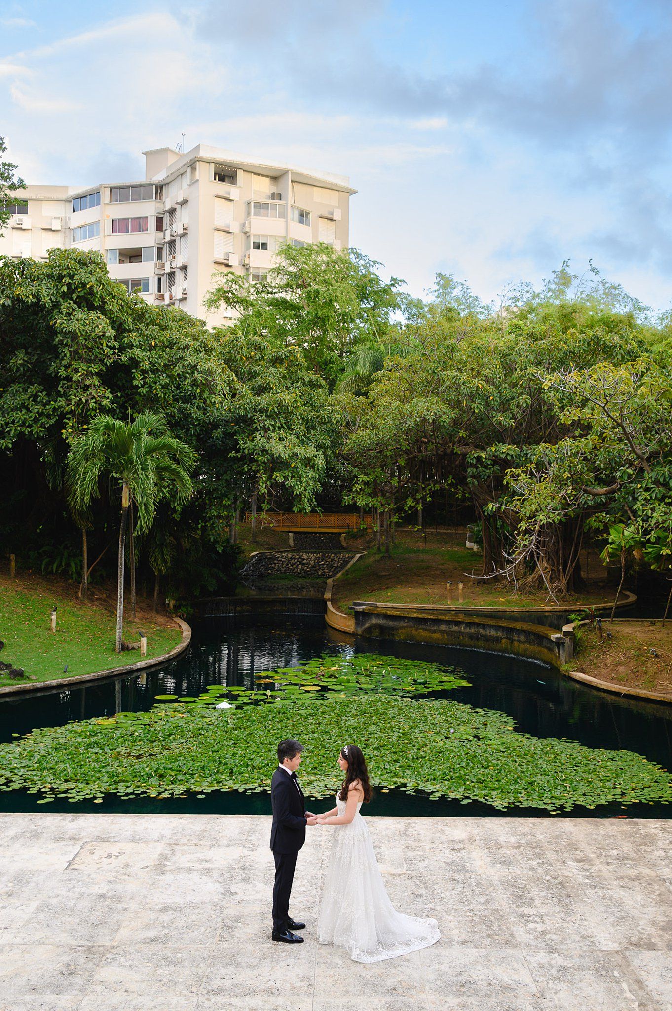 Bride and groom stand facing each other on a terrace beside a lily pond surrounded by lush greenery and buildings.