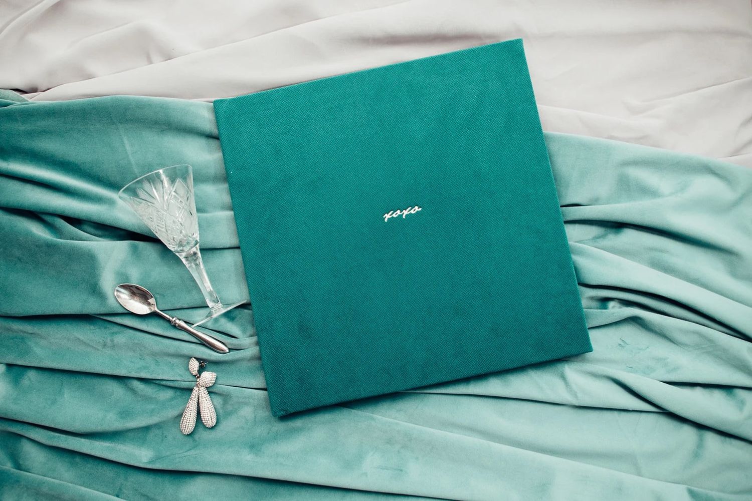 Teal velvet wedding photo album with silver spoons and crystal glass on matching fabric.