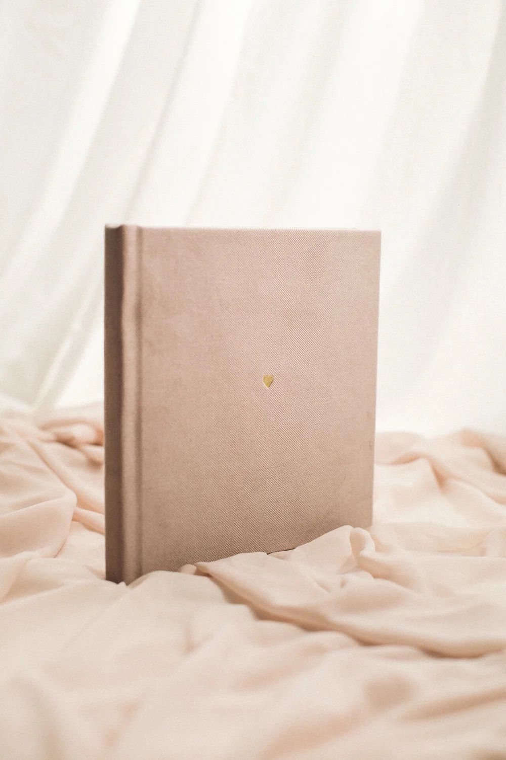 Elegant blush pink hardcover photo album with gold heart detail, displayed on soft fabric with white curtain background.