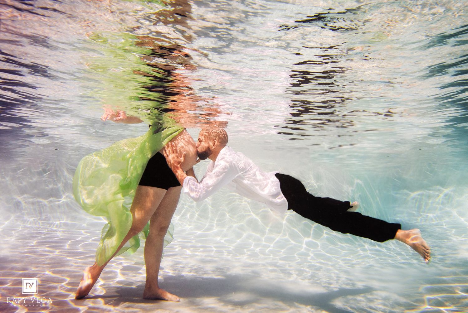 Couple sharing a romantic underwater kiss, woman in flowing green dress and man in white shirt and black pants.