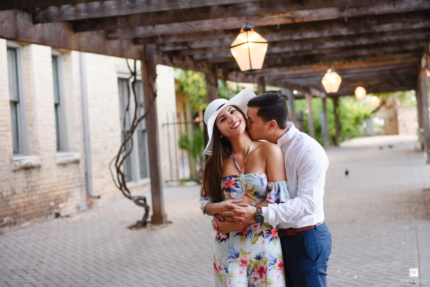 Couple sharing a romantic kiss under vintage street lamps in a charming urban courtyard, woman in floral dress and white hat.