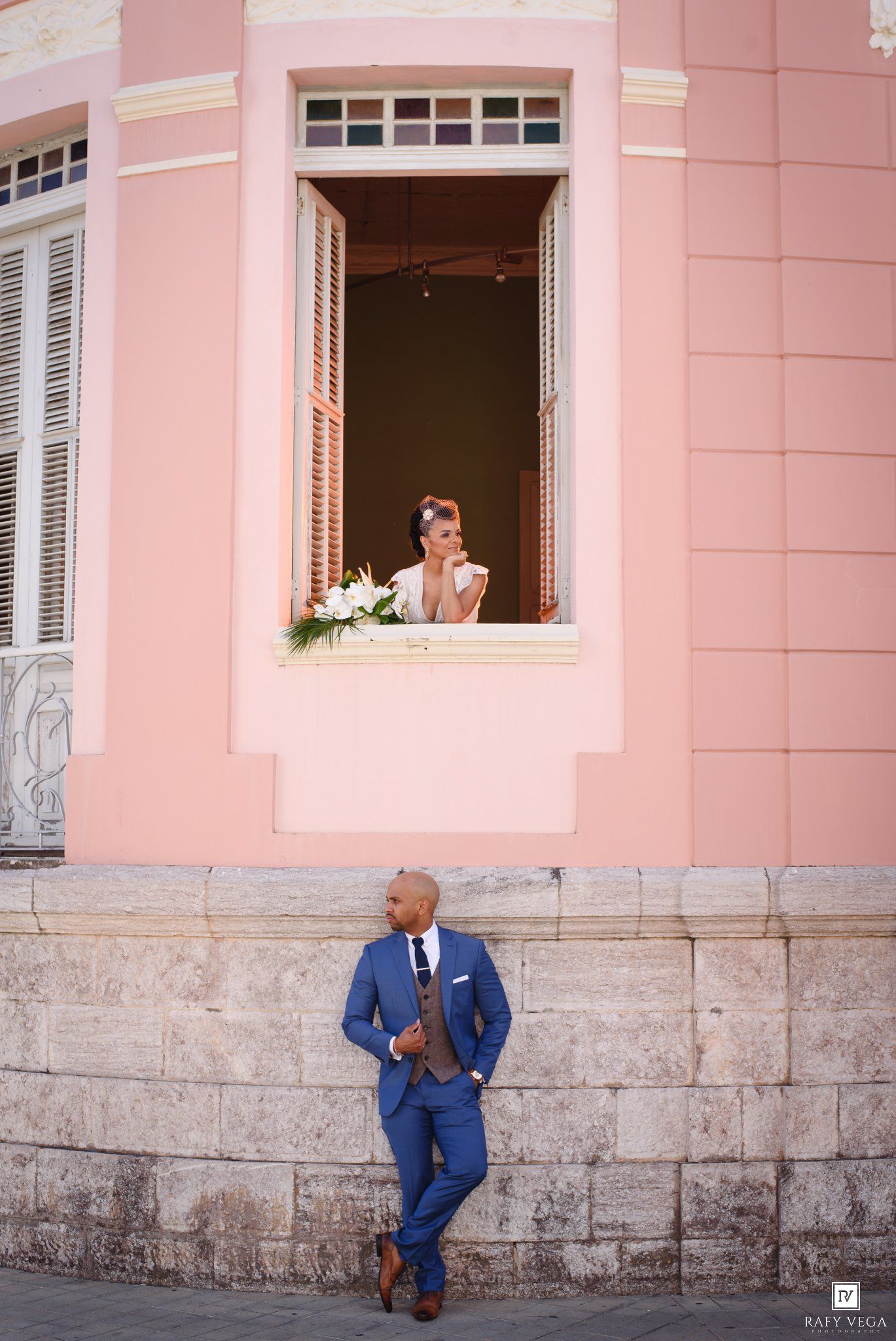 Groom in blue suit leaning against pink building wall while bride looks out from window above.