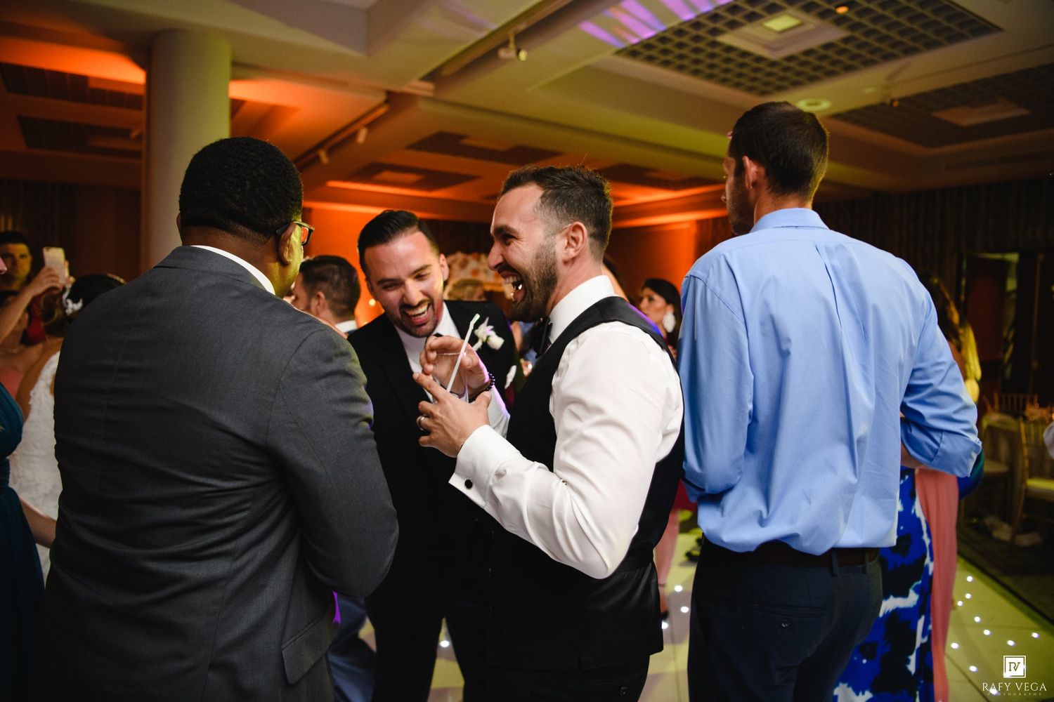 Groom laughing with groomsmen on wedding reception dance floor in elegant ballroom.