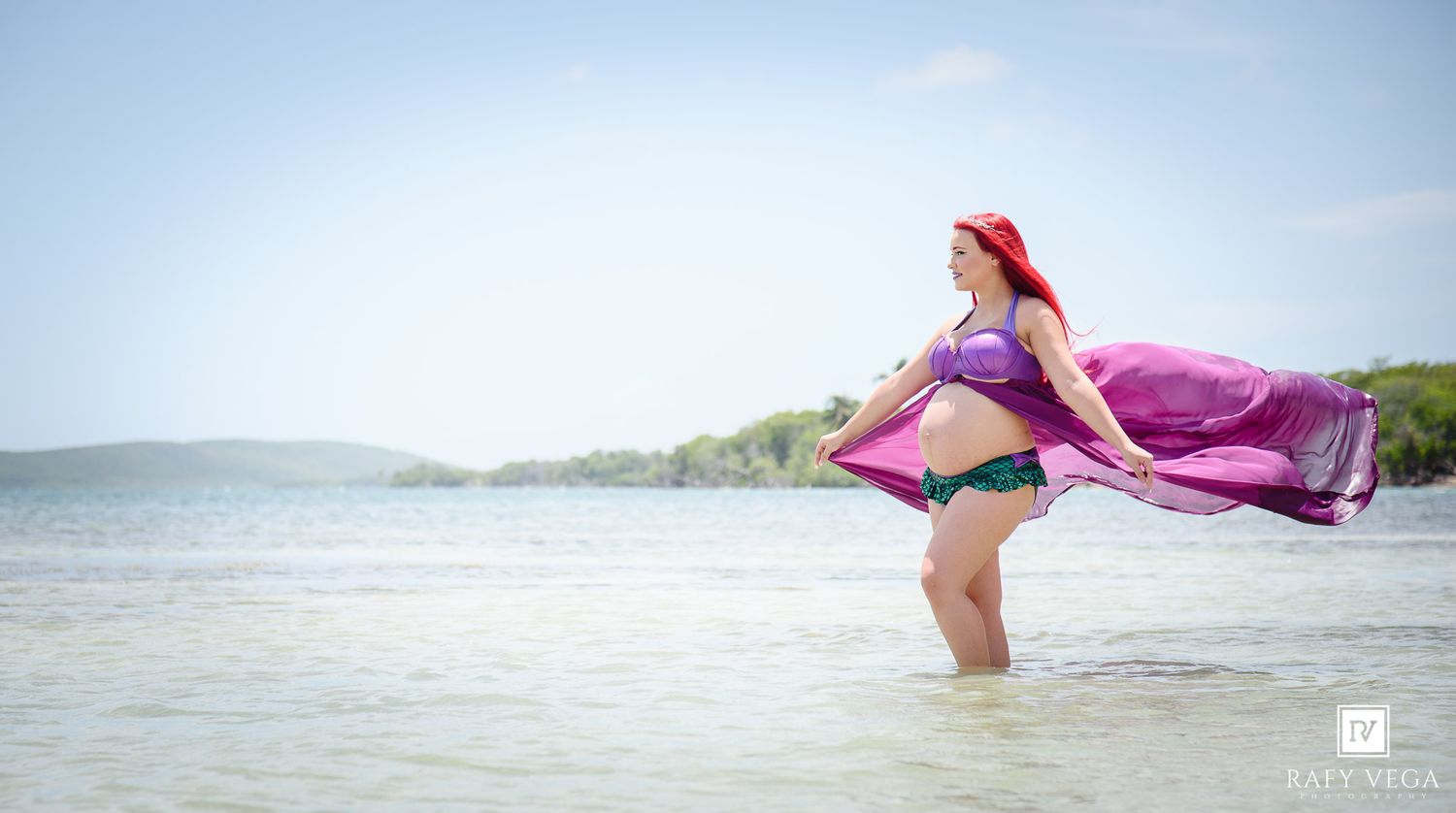 Woman in mermaid costume with red wig and purple fabric poses in shallow ocean water on a sunny beach day.