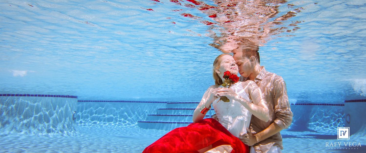 Romantic couple embracing underwater in a pool, woman in red skirt holding a rose, man in white shirt.