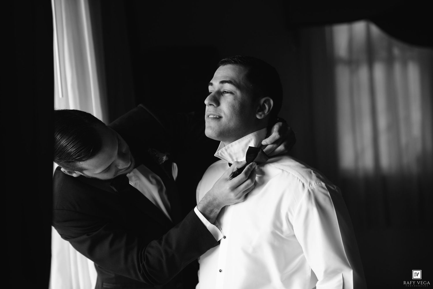 A groomsman adjusts the bow tie of a groom in a white dress shirt before a wedding ceremony.