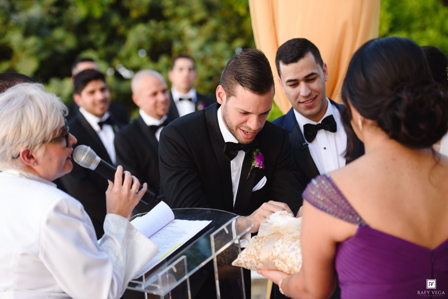 Groom in black tuxedo signing wedding documents at outdoor ceremony with groomsmen and officiant.