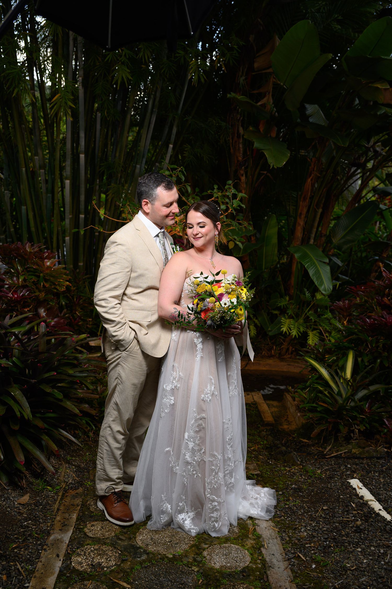 Bride and groom pose together in lush tropical garden, bride holding colorful bouquet in white lace gown, groom in tan suit.