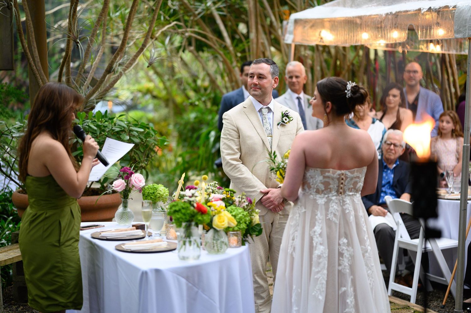 Outdoor wedding ceremony with bride and groom holding hands as officiant reads vows, surrounded by lush greenery and guests.