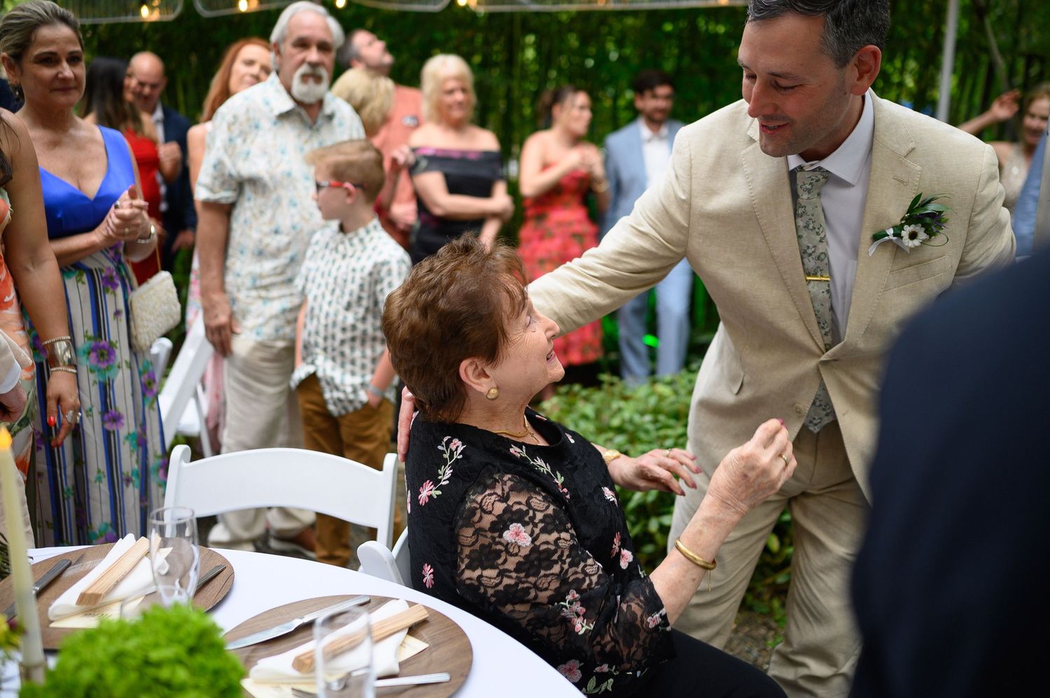 A groom in a beige suit greets a seated elderly woman at an outdoor wedding reception with guests in the background.