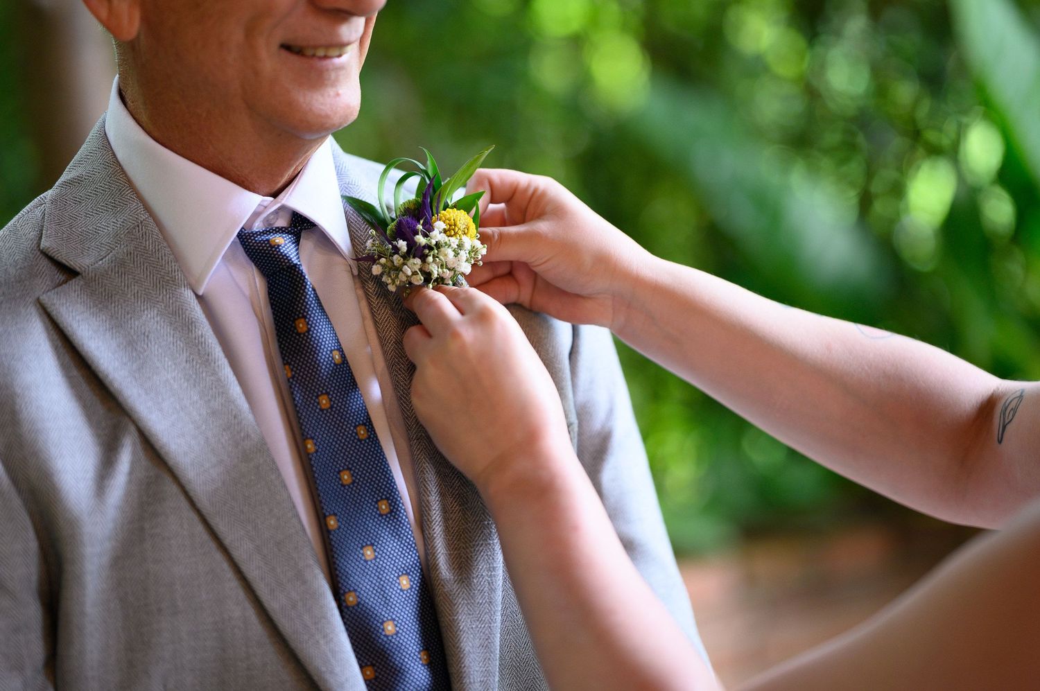 Hands pinning a floral boutonniere on a man in a gray suit with a blue polka dot tie at a wedding.