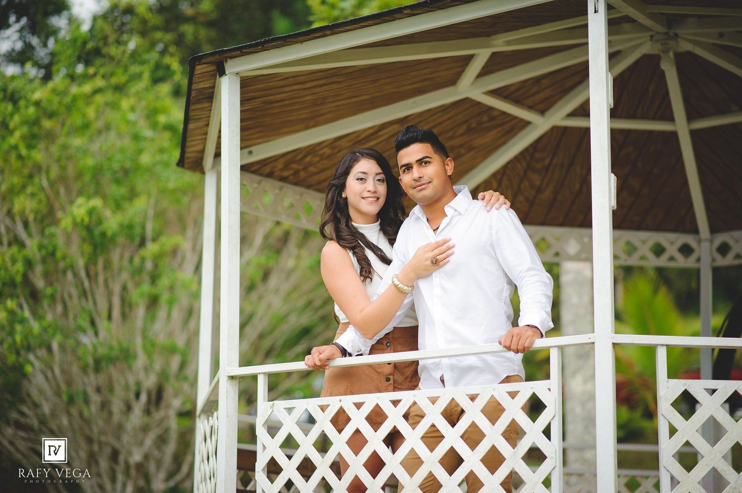 Couple in white outfits posing in a white gazebo surrounded by lush green trees in a romantic outdoor setting.