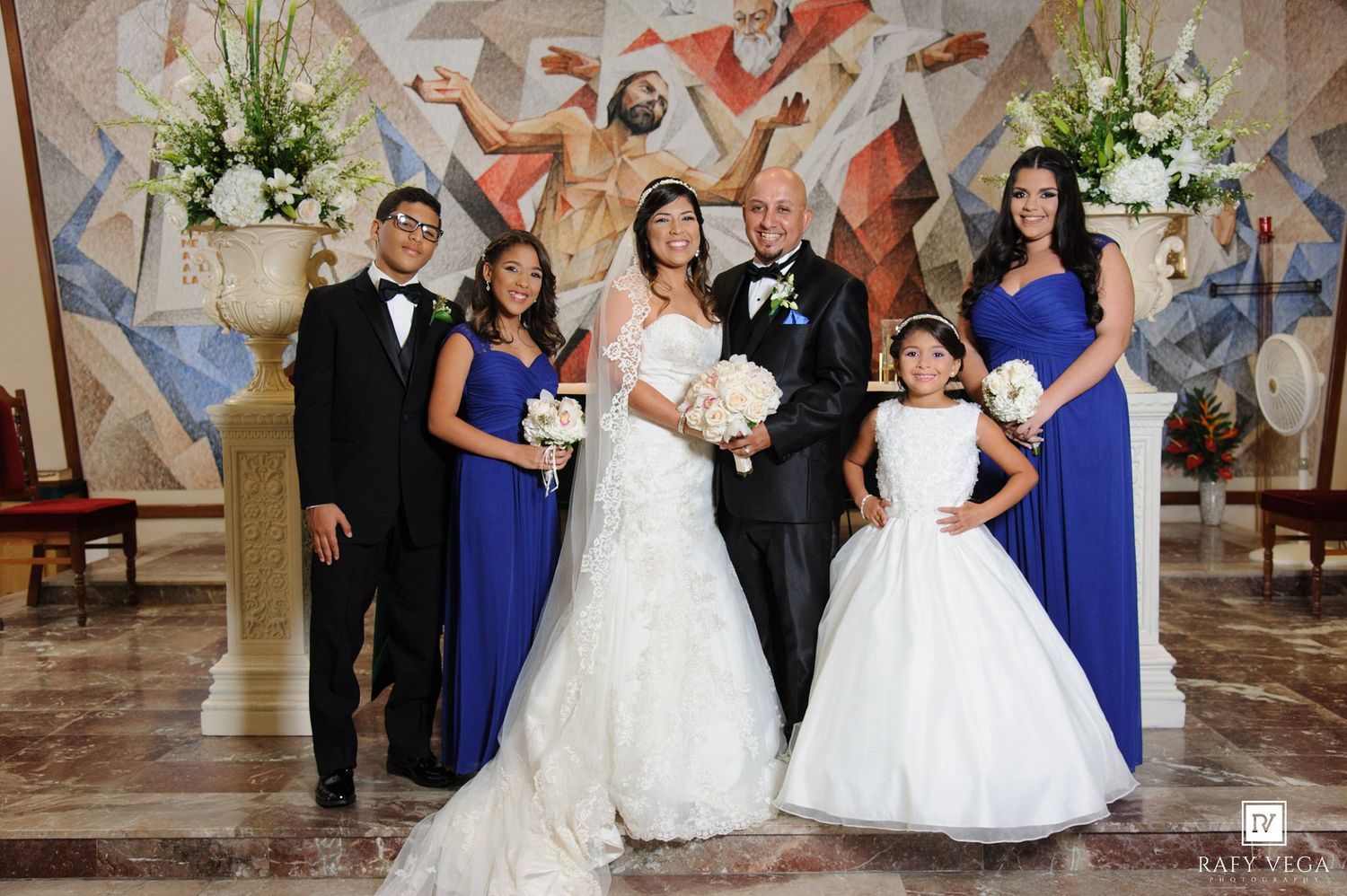 Wedding party poses in church with bride in white gown, groom, bridesmaids in blue, groomsman, and flower girl.