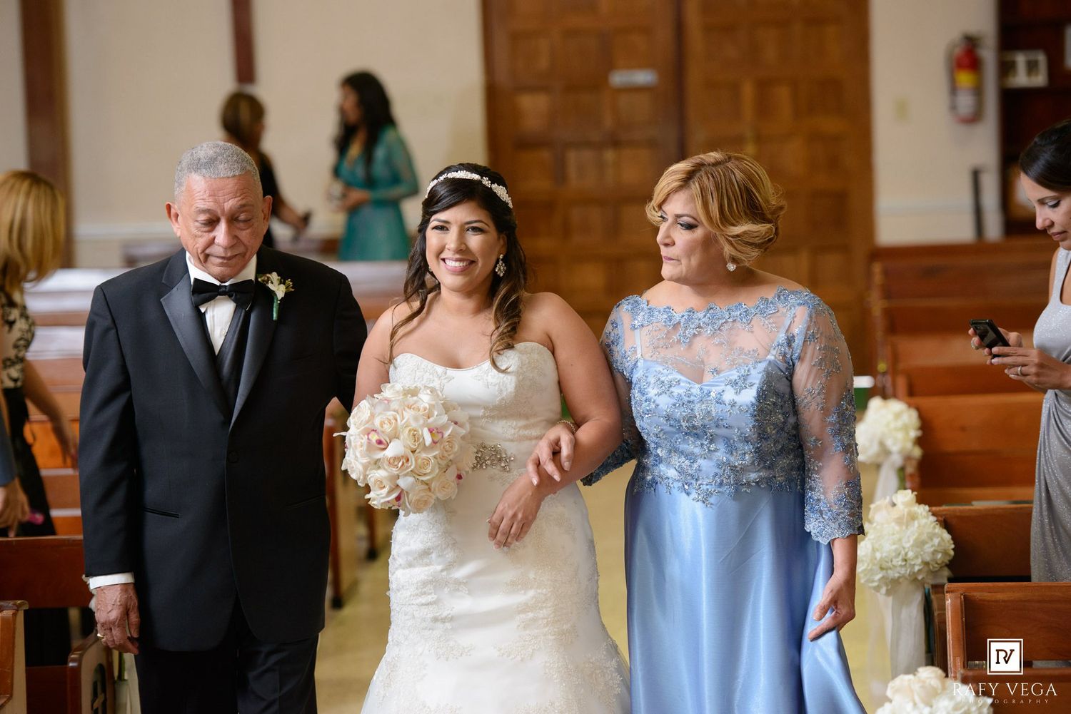 Bride walks down church aisle escorted by parents, wearing strapless gown, holding white bouquet, mother in blue lace dress.