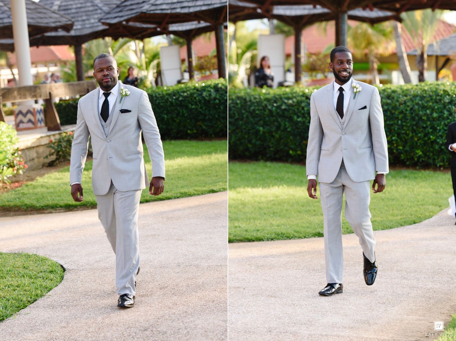 A well-dressed man in a light gray suit and black tie walks outdoors at a wedding venue surrounded by lush greenery.