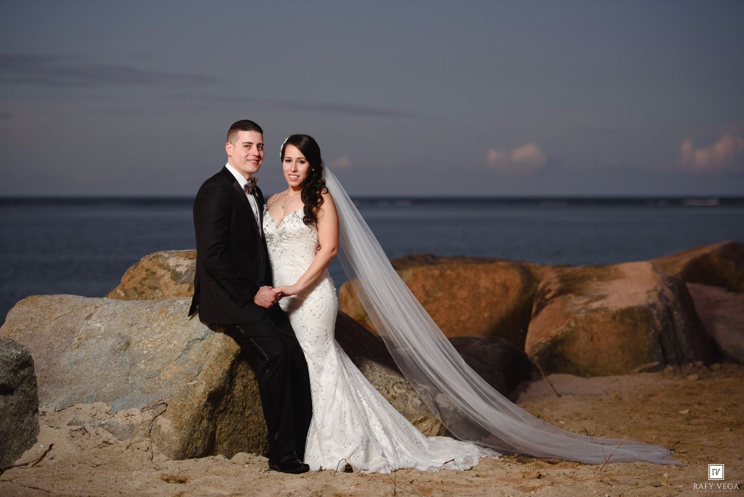 Bride in mermaid gown with cathedral veil and groom in tuxedo pose on rocky beach at sunset.